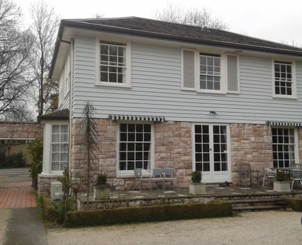 Exterior view of a two-storey traditional-style house with pale horizontal weatherboard siding on the upper level, coursed light stone cladding on the lower level, white multi-pane windows with shutters, French doors, striped awnings, and a low landscaped terrace. The house has a shallow hipped roof and a formal, restrained facade.