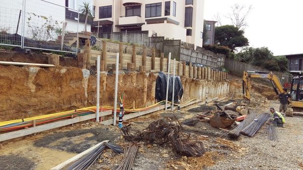 A residential construction site on a sloped suburban lot, showing exposed earth excavation, temporary retaining and boundary structures, reinforcing steel, utility conduits, construction materials, and heavy machinery. A multi-storey modern house is visible above the excavation.