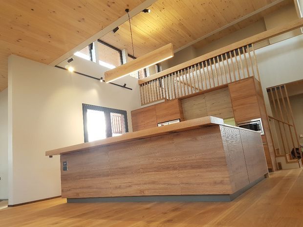 A warm, open-plan kitchen interior with extensive timber finishes. The room features a large timber island with integrated cabinetry and a raised benchtop, timber flooring, a timber-lined ceiling, a long linear pendant light, and a timber balustrade overlooking a mezzanine or upper-level landing. High-level glazing and exposed open volume create an airy, loft-like space.