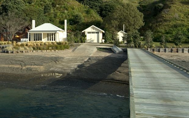 A small waterfront residential compound viewed from the water, with light-coloured single-storey buildings set against a steep, densely vegetated hillside. The main cottage has a low hipped roof, verandah-like glazing and pale vertical cladding, with two smaller outbuildings behind it. A long timber jetty extends from the shore into the foreground water, while gravel landscaping and rock edging define the shoreline.