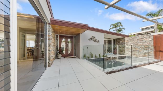 Contemporary residential courtyard viewed from an outdoor paved terrace, featuring a small glass-edged swimming pool, large-format pale tiled paving, stone-clad walls, timber entry doors, broad glazing, and a covered outdoor area. The low-pitched rooflines, clean geometric forms, and warm timber and stone accents create a refined modern exterior.
