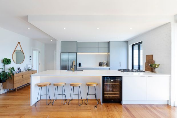Bright, open-plan contemporary kitchen with a large white island and four timber-topped stools, pale grey full-height cabinetry, integrated stainless-steel refrigerator, built-in oven, gas cooktop, and warm timber flooring. A white-painted brick feature wall and black-framed windows add contrast, while a timber sideboard and round wall mirror sit in the adjoining area.