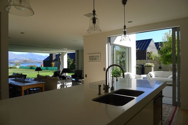 A bright, open-plan contemporary kitchen viewed from behind a large island. The island has a pale smooth benchtop, dark double sink, and curved dark tap, with pendant lights overhead. Beyond it is an open dining and living area with extensive glazing and views toward a landscaped outdoor area, lake, and mountains. A patio with outdoor seating is visible through the doors.