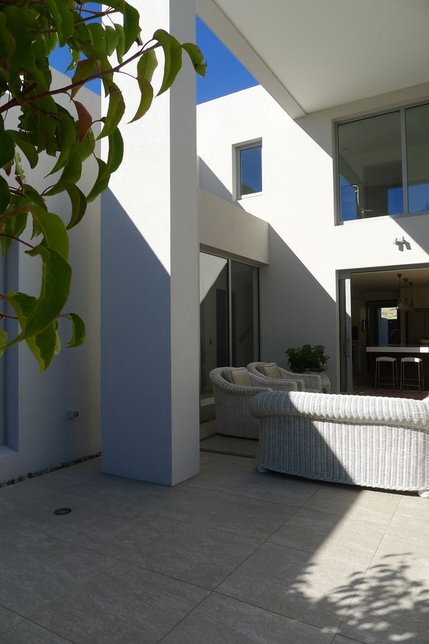 A bright modern courtyard or outdoor entertaining area enclosed by white rendered walls and strong geometric columns. Large grey porcelain or stone-look tiles form the floor, with woven light-grey outdoor lounge chairs and a matching sofa in the foreground. Glazed doors and windows connect the courtyard to an interior kitchen or bar area. Deep angular shadows, blue sky, and leafy planting create a crisp, sunlit atmosphere.