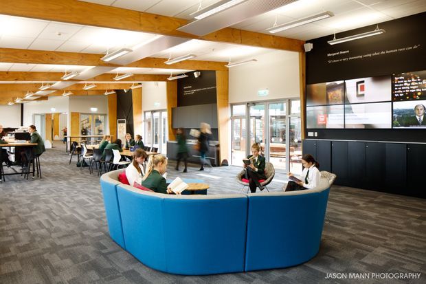 A bright contemporary open-plan commercial interior, likely a library, learning commons, or university student lounge. The space features a large curved blue upholstered seating booth in the foreground, scattered tables and chairs, timber structural beams, grey carpet tiles, extensive digital display screens, and suspended linear lighting. People are using the space for reading, studying, and conversation.