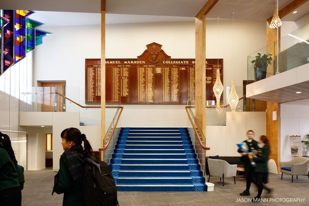 A bright, double-height institutional foyer with a wide blue-carpeted staircase, timber posts and handrails, a large heritage honor board, colorful stained-glass artwork, pendant lights, upper-level glazing, and seating areas. People are walking through the space.