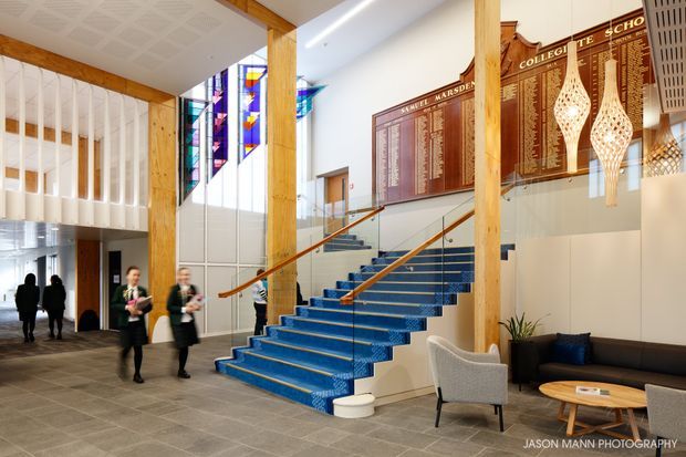 A bright institutional interior lobby featuring a dramatic blue-carpeted staircase with timber handrails, exposed timber columns, stained-glass windows, wall-mounted honor boards, and a small seating area. The space has a generous vertical volume, patterned acoustic ceiling panels, and polished contemporary school or college foyer detailing.