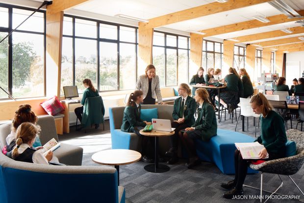 Bright, open-plan educational common area with students working and socialising across informal lounge seating, tables, and high communal benches. The space features extensive black-framed glazing, exposed laminated timber beams and columns, acoustic ceiling panels, carpet flooring, and a contemporary school or campus interior.