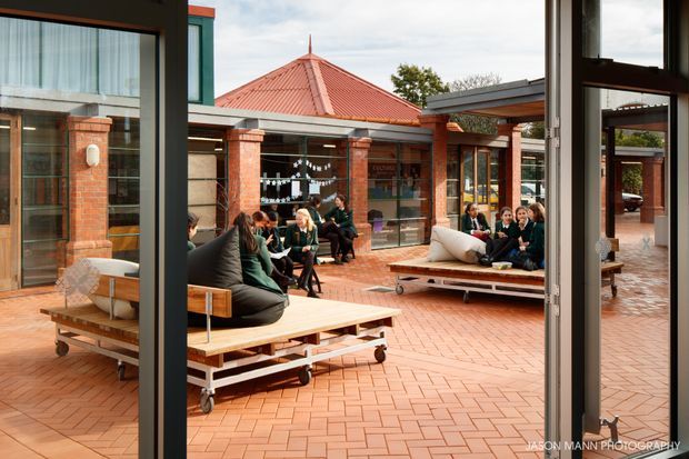 A view from inside a glazed building looking onto a spacious brick-paved courtyard. The courtyard is framed by red-brick low-rise buildings with large windows and a distinctive terracotta hipped roof. Students or visitors gather around movable timber-and-metal seating platforms, creating an active communal outdoor space. Dark window and door frames contrast with the warm brickwork.