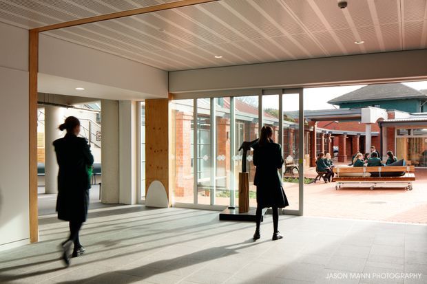 A bright contemporary institutional interior opens onto a brick-paved courtyard. The space features large glazed sliding doors, exposed timber posts, pale tiled flooring, a white perforated linear ceiling, and strong sunlight casting long shadows. People are visible inside and in the courtyard beyond.