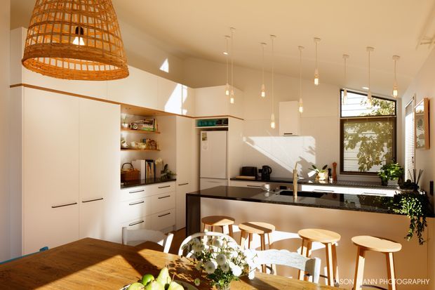 Bright, warm contemporary kitchen and dining area with white full-height cabinetry, open shelving, a central island with dark benchtop, timber bar stools, pendant lighting, and a large timber dining table. A woven rattan pendant adds a natural feature above the dining zone, while sunlight enters through the windows and highlights the clean, welcoming interior.