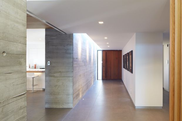 A bright modern residential hallway viewed toward a solid timber entry door. The corridor features a long exposed concrete wall, smooth light walls, large neutral floor tiles, recessed ceiling downlights, and a timber-framed opening at the foreground. A glimpse of a minimalist kitchen or bar area with a stool is visible to the left, while framed artwork lines the right wall.