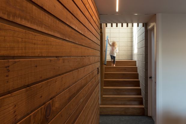 A warm, narrow interior stairway with broad timber-clad walls and matching timber stair treads. The stairs lead upward toward a bright glazed landing, with a slender linear ceiling light and a contrasting grey masonry wall. A doorway opens on the right.