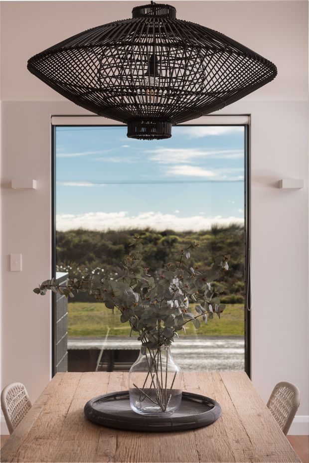 Interior dining area with a rustic light-wood dining table, woven white chairs, a large black woven pendant light, and a glass vase of foliage. A full-height window frames a bright coastal or rural landscape beyond, while white walls and minimal wall lighting create a relaxed, natural atmosphere.