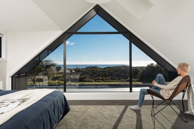 A bright contemporary bedroom with a dramatic triangular floor-to-ceiling window and black-framed glazing overlooking a coastal landscape. The room includes a partially visible bed with dark blue bedding, pale walls and ceiling, grey carpet, and a simple lounge chair occupied by a person. The angular window follows the pitched roof form and creates a strong architectural focal point.