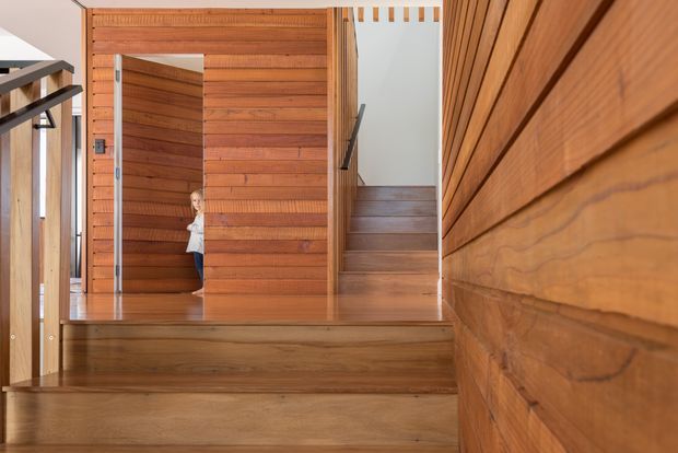 Interior view of a warm timber-lined entry and stair hall. Broad timber-clad walls, doors, stair treads and flooring create a cohesive built-in wood interior, with a person partially visible through a doorway in the background and a simple dark handrail along the stairs.