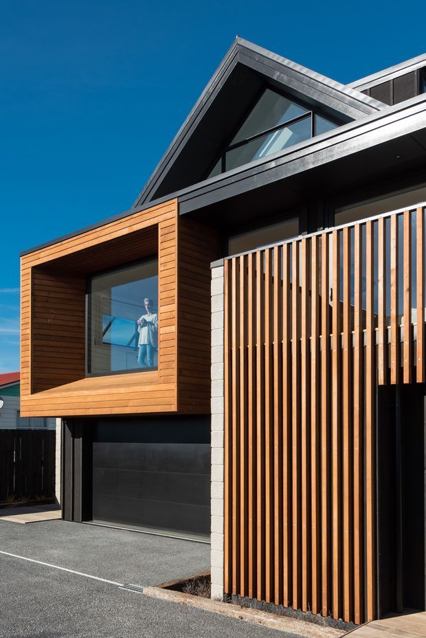 Exterior view of a contemporary multi-level residential building with a steep gabled roof, dark cladding, warm timber-clad projecting window bay, large glazing, and a vertical timber slat privacy screen. A garage door and paved driveway are visible at ground level.