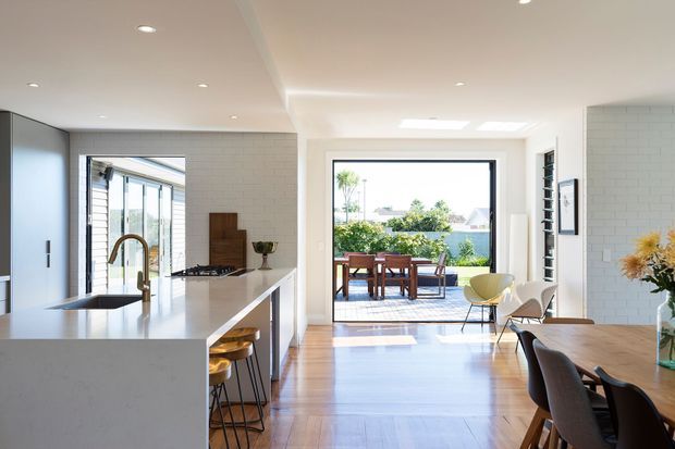 Bright open-plan kitchen and dining area with a large white island, integrated cooktop, timber flooring, and contemporary dining furniture. Full-height glazing opens to an outdoor terrace with water visible beyond, while white brick-style wall tiles and skylights enhance the airy interior.