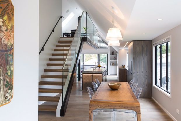Bright contemporary open-plan interior featuring a timber dining table and transparent chairs in the foreground, a compact kitchen beyond, and a sculptural open-riser staircase with timber treads, black handrail, and glass balustrade. Floor-to-ceiling timber cabinetry lines the right wall, while large windows and a sloped ceiling bring in abundant daylight.