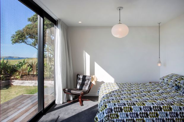 Bright, compact bedroom with a large black-framed glazed sliding door opening to a timber deck and landscaped coastal-looking garden. The room has white walls, grey carpet, sheer curtains, a patterned bedspread, a leather lounge chair, and simple pendant lighting. Strong sunlight creates a calm, airy interior with clear indoor-outdoor connection.