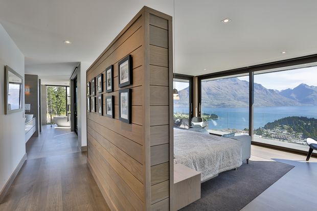 A bright modern interior showing a hallway divided from a bedroom by a full-height horizontal timber feature wall displaying framed artwork. The bedroom has floor-to-ceiling glazing with expansive views over a blue lake, mountains and a settled shoreline. Visible finishes include warm timber flooring and wall panelling, white walls and ceiling, dark window frames, and a neutral upholstered bed with a grey rug.