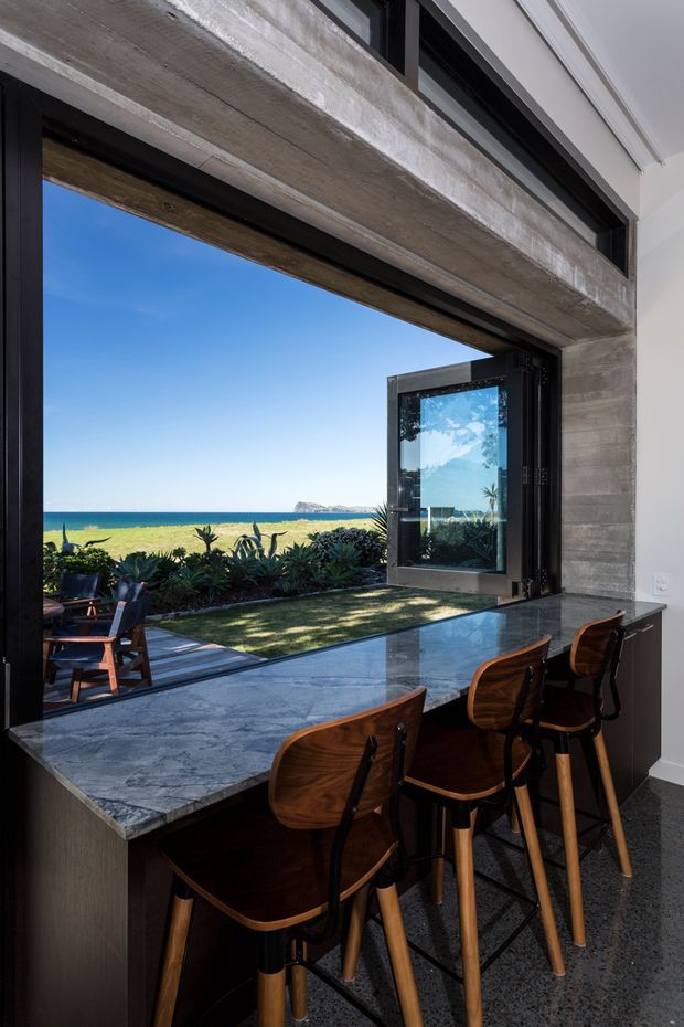 A contemporary kitchen or entertaining space with a dark stone peninsula and timber bar stools positioned beside a large open corner window. The opening frames a lawn, coastal planting, sandy beach, turquoise water, and a distant headland. Exposed concrete surrounds the glazing, while black-framed windows and doors create a strong indoor–outdoor connection. The image is bright, coastal, and refined, with a clear view toward the ocean.