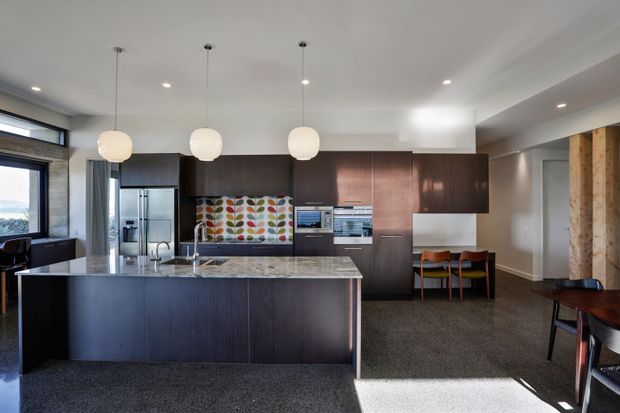 A contemporary open-plan kitchen dominated by a large dark timber island with a pale stone benchtop. Full-height dark cabinetry incorporates stainless-steel refrigeration and built-in appliances, while a colourful patterned tiled splashback adds visual interest. Three spherical pendant lights hang above the island, with a compact dining area and expansive glazing visible around the edges.