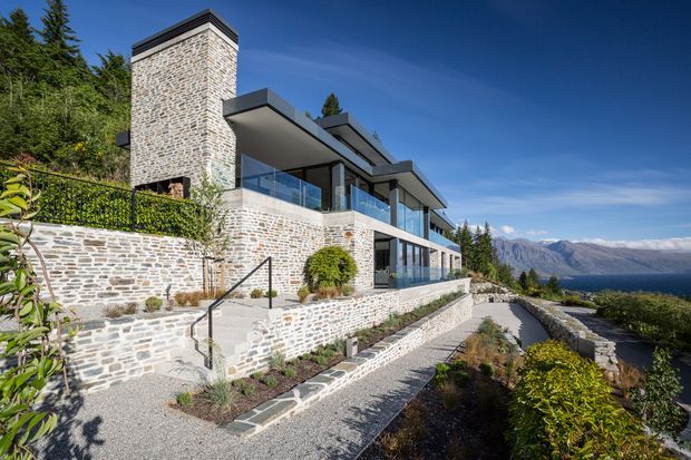 Exterior view of a contemporary multi-level residence built into a landscaped hillside. The home features extensive pale stone masonry, dark flat roof planes, large glazed openings with glass balustrades, terraces, retaining walls, and a long stepped pathway. A mountainous lakeside landscape is visible beyond the property.