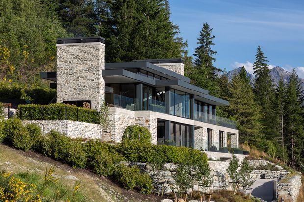 Contemporary multi-level mountain residence set into a landscaped sloping site, with extensive floor-to-ceiling glazing, dark flat roof planes, and prominent light stone masonry chimneys and walls. Terraced stone retaining walls, hedges, mature conifers, and distant alpine peaks surround the home.