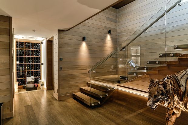 A warm, sophisticated interior hallway with dark timber flooring and horizontal timber wall panelling. A floating timber staircase with a clear glass balustrade rises along the right side, illuminated by recessed and wall-mounted lighting. At the rear is a temperature-controlled wine display or cellar with extensive bottle storage, while sculptural timber decor occupies the foreground.