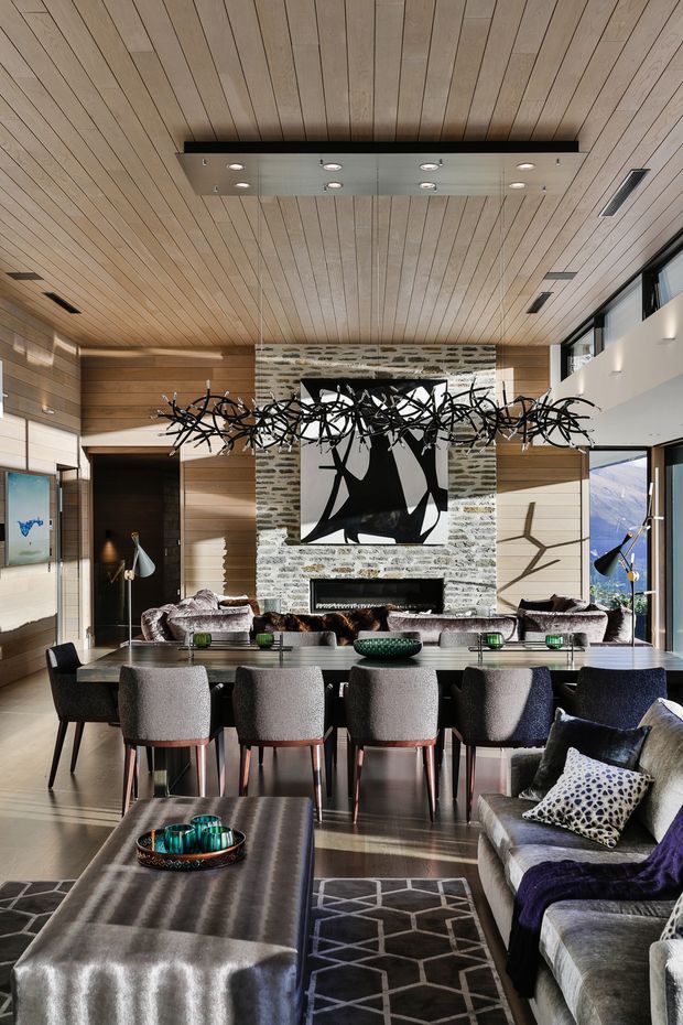 A sophisticated open-plan living and dining room with a long dining table, upholstered dining chairs, a large sectional sofa, and a fireplace wall clad in horizontally coursed stone. Warm timber wall and ceiling panelling create a rich architectural backdrop, while expansive glazing brings in abundant daylight and views. A dramatic sculptural black pendant spans the dining area, complemented by contemporary artwork, layered rugs, and metallic accents.