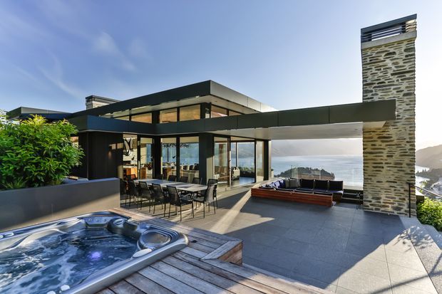 Contemporary multi-level hillside residence with expansive glazing, dark metal cladding, a prominent natural stone chimney, and an open terrace overlooking a coastal or mountainous landscape. The outdoor area includes a spa pool, timber decking, dining setting, and built-in outdoor seating.