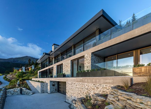 A contemporary multi-level hillside residence with strong horizontal lines, extensive floor-to-ceiling glazing, glass balustrades, projecting flat roof planes, and textured natural stone masonry walls. The home is integrated into a landscaped sloping site with terraced retaining walls and mountain views.