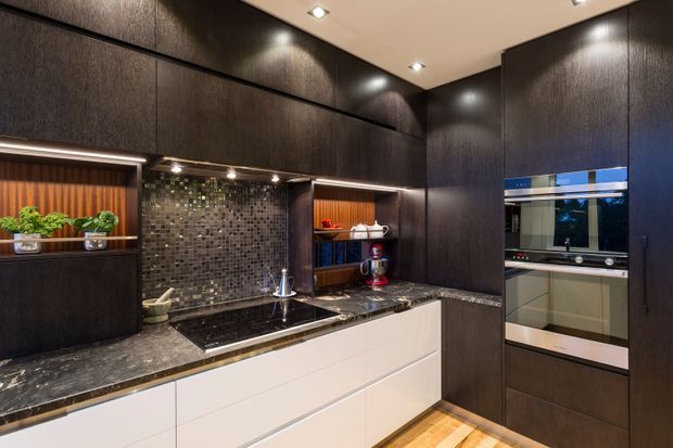 A compact, sophisticated contemporary kitchen with dark full-height cabinetry, a white island with integrated sink, dark stone benchtops, and a small dark mosaic tile splashback. Built-in ovens and open display shelving are integrated into the cabinetry, with warm timber flooring and recessed ceiling lighting creating a refined, moody atmosphere.