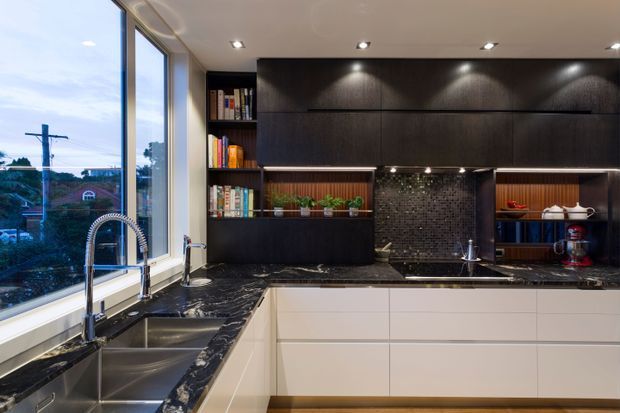A contemporary kitchen with extensive dark built-in cabinetry, white lower drawers, black marble-look benchtops, a textured dark mosaic splashback, stainless-steel double sink and tap, open timber display shelving, and integrated warm lighting. Large corner windows provide views to the outdoors.