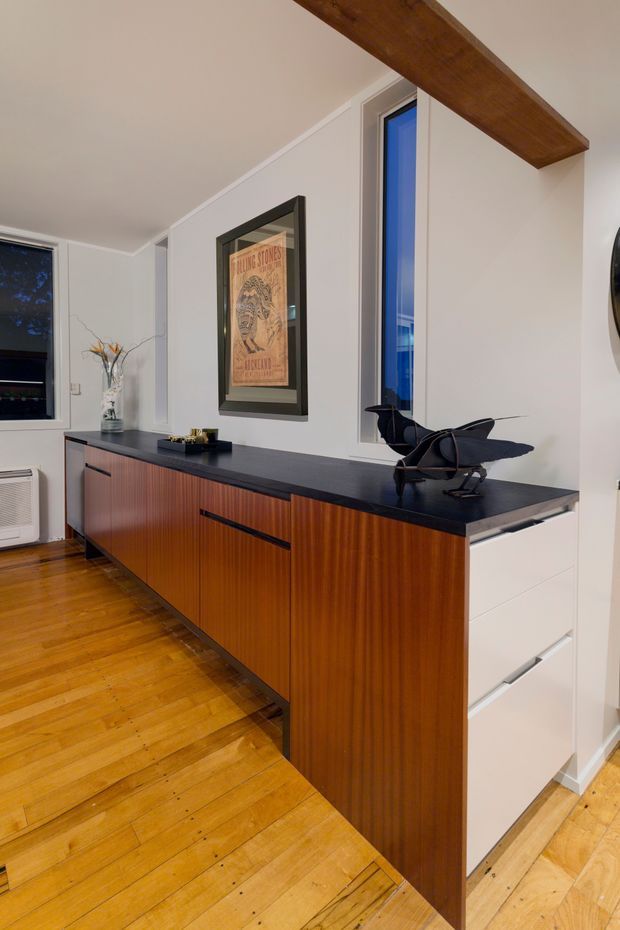A refined interior hallway or living-area storage zone featuring a long built-in sideboard with warm vertical-grain timber cabinetry, a dark stone-like benchtop, and integrated white drawers at one end. White walls, timber flooring, a framed vintage poster, narrow windows, and an exposed dark timber ceiling beam create a warm mid-century-inspired character.