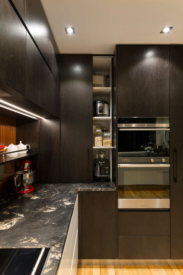 A compact, sophisticated galley kitchen with full-height dark timber cabinetry, integrated double wall ovens, open appliance shelving, and a dramatic dark stone benchtop with light veining. Warm under-cabinet lighting and recessed ceiling spotlights illuminate the space, while a timber floor adds warmth and contrast.