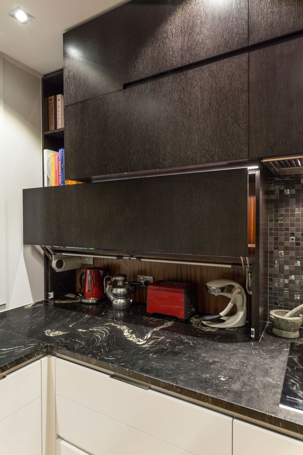 Close-up view of a compact, dark-toned kitchen work area with floor-to-ceiling dark wood-grain cabinetry, integrated lift-up storage, open shelving with books, and a black veined stone benchtop. A metallic mosaic tile splashback sits behind a concealed cooking or appliance zone, with a kettle, toaster, stand mixer and other small appliances arranged on the counter. Recessed ceiling lighting and pale lower cabinetry provide contrast.
