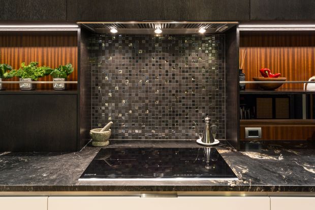 A dramatic contemporary kitchen cooktop area with a black stone benchtop, dark cabinetry, warm timber-lined open shelving, and a full-height reflective dark mosaic tile splashback. Integrated lighting highlights the cooktop and display niches, while small plants, cookware, and decorative objects add warmth.