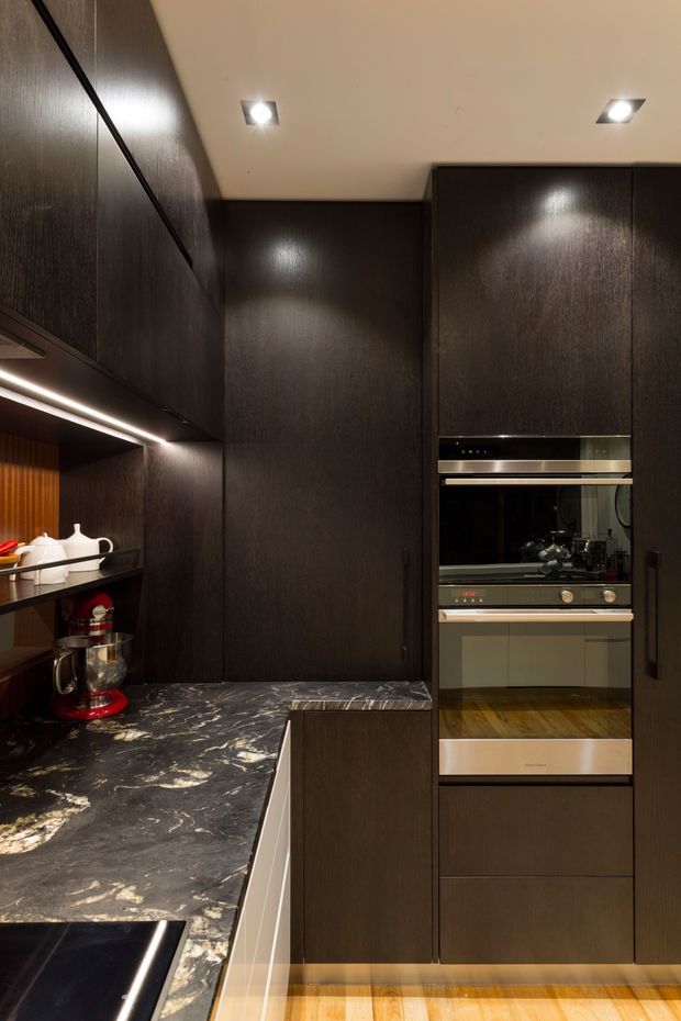 A compact, sophisticated kitchen featuring dark wood-grain full-height cabinetry, integrated double ovens, a dark veined stone benchtop, under-cabinet lighting, recessed ceiling spotlights, and timber flooring. A red stand mixer and white serving pieces provide small contrasting accents.