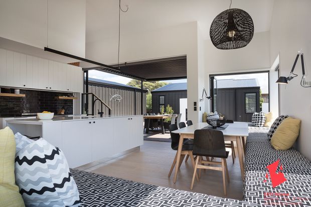 Bright open-plan kitchen and dining area with a white island, dark tiled splashback, timber flooring, and a built-in patterned bench seat. The space opens to a covered outdoor courtyard through large glazed doors, with a dining table, pendant lighting, and contemporary black-and-white furnishings.