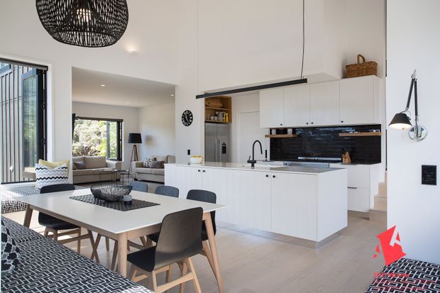 Bright open-plan kitchen and dining area with a white island, extensive white cabinetry, glossy dark backsplash, and integrated appliances. A light timber dining table with dark upholstered chairs occupies the foreground, while the living area is visible beyond. The space features pendant lighting, a wall-mounted task lamp, pale timber-look flooring, and a clean contemporary fit-out.