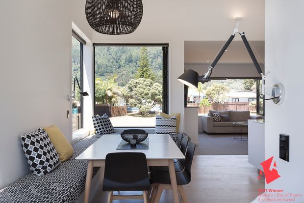 Bright contemporary dining area with a white rectangular dining table, black chairs, patterned upholstered bench seating, and a large woven pendant light. The room opens directly into a living room with a grey sofa and another sculptural pendant light. Large windows and glazing provide views of landscaped greenery and neighbouring homes. Light timber flooring, white walls, and black accents create a crisp, graphic interior.