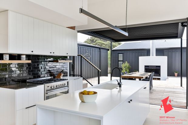 Bright contemporary kitchen opening onto a covered outdoor dining deck. The room features white flat-panel cabinetry, a large white island with integrated sink, black reflective subway-tile splashback, stainless-steel range and appliances, and slim black linear pendant lighting. Beyond the kitchen is a timber deck with an outdoor dining table and a freestanding white fireplace against dark vertical cladding.