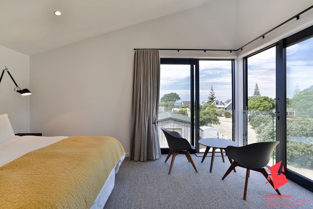 Bright contemporary bedroom with a sloped white ceiling, neutral walls, carpeted floor, and a bed dressed with a mustard-yellow quilt. A glazed corner window wall with dark frames opens to a scenic suburban view, while a small round table and two dark lounge chairs form a sitting area beside the windows. Minimal black track lighting and a slender floor lamp complete the room.