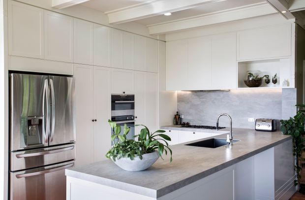 Bright, refined contemporary kitchen with extensive white flat-panel cabinetry, a large central island with a grey stone-look benchtop, integrated sink and gooseneck tap, stainless-steel French-door refrigerator, built-in oven, and a matching grey splashback. Recessed ceiling lighting and decorative ceiling beams add architectural detail, while potted greenery provides a natural accent.