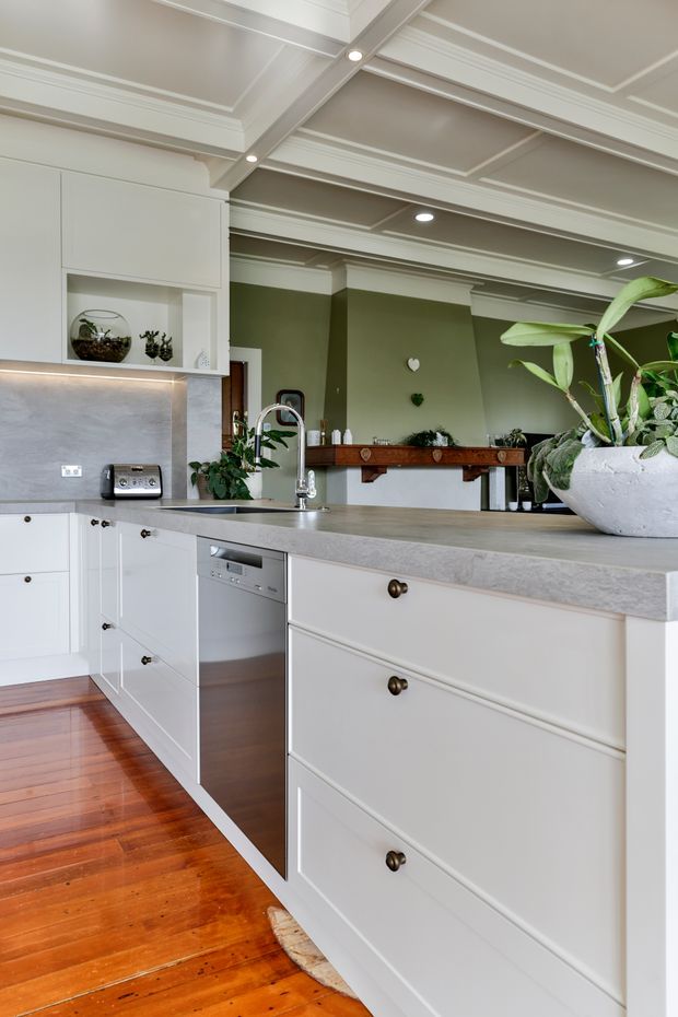 A bright, traditional-style kitchen with white Shaker cabinetry, a large grey stone island benchtop, integrated dishwasher, stainless-steel tapware, and a grey stone splashback. The room features polished warm timber flooring, an olive-green feature wall, decorative timber wall shelving, and a detailed coffered ceiling with recessed lighting. Potted plants add an organic accent.