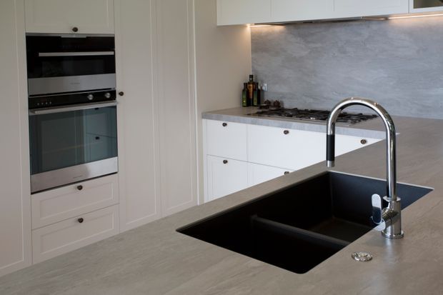 A bright, refined kitchen with white Shaker-style cabinetry, integrated stainless-steel ovens, a large grey stone-look island benchtop, deep black undermount sink and polished chrome gooseneck tap. A matching grey splashback and under-cabinet lighting complete the clean, contemporary fit-out.