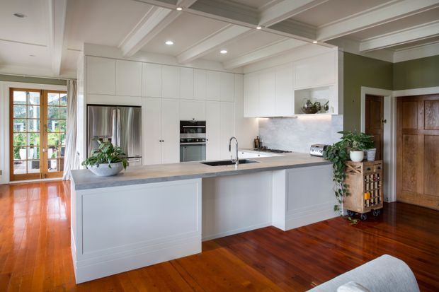 A bright, spacious kitchen with a large central island, white full-height cabinetry, integrated appliances, a stainless-steel refrigerator, and a light stone benchtop. The room features warm polished timber flooring, a coffered white ceiling with recessed lighting, French doors opening to greenery, and a dark olive-green adjacent wall with traditional timber doors.
