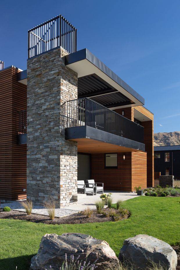 Exterior architectural photograph of a modern multi-level residence with a strong vertical stone-clad tower, dark metal balustrades, flat projecting roof planes, and warm horizontal timber cladding. The building overlooks a landscaped lawn with ornamental grasses and large natural rocks, with rugged hills visible in the background.